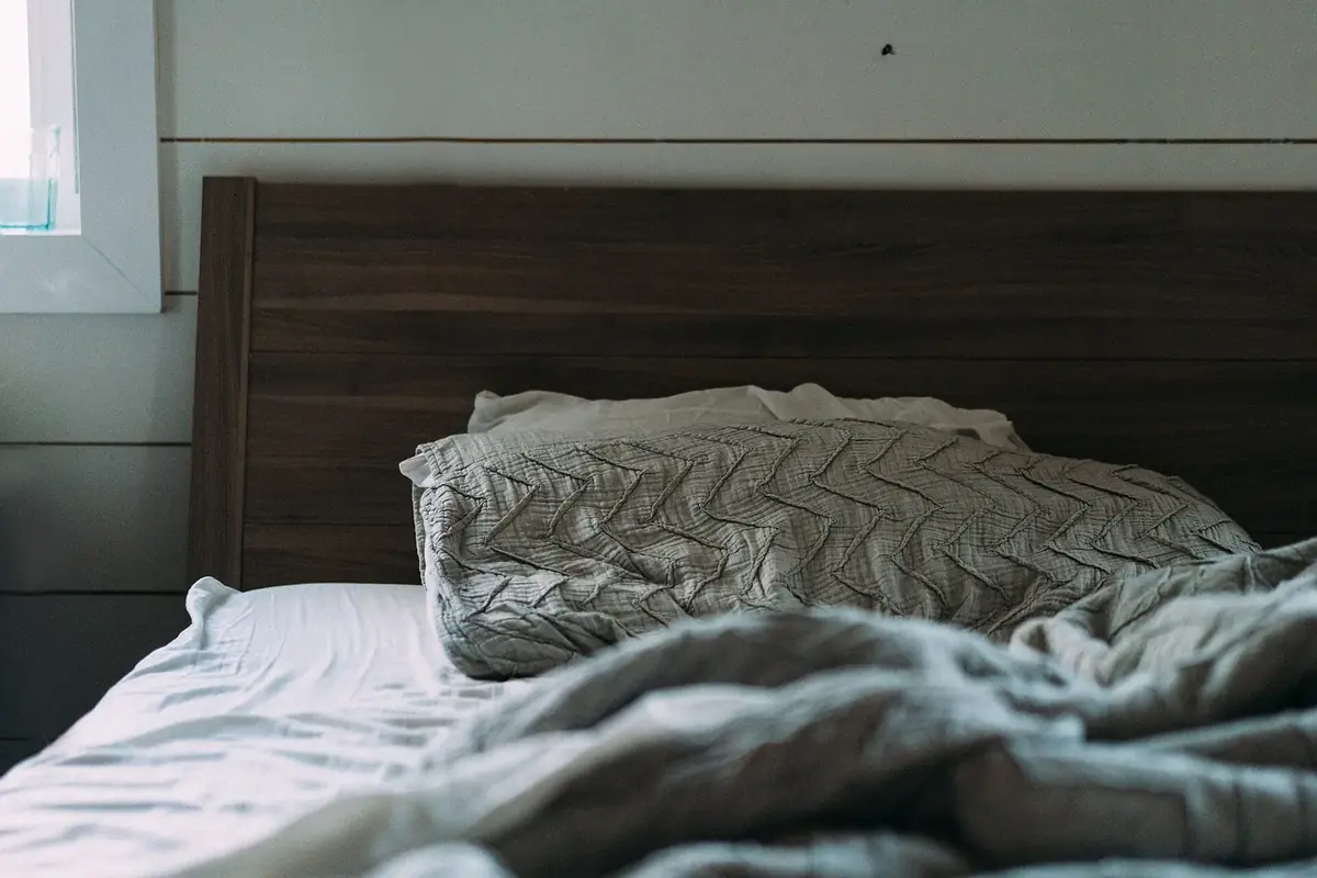 An unmade bed with a wooden headboard, a stitched grey pillow and rumpled linen against a white plank wall
