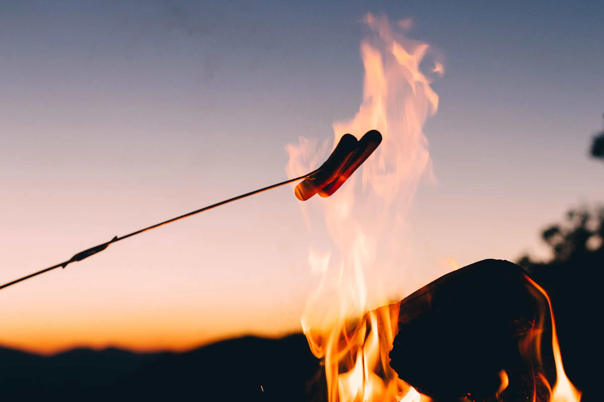 Two sausages on a long roasting fork held over the flames of a campfire, against a dusk sky fading from blue to orange above dark hills